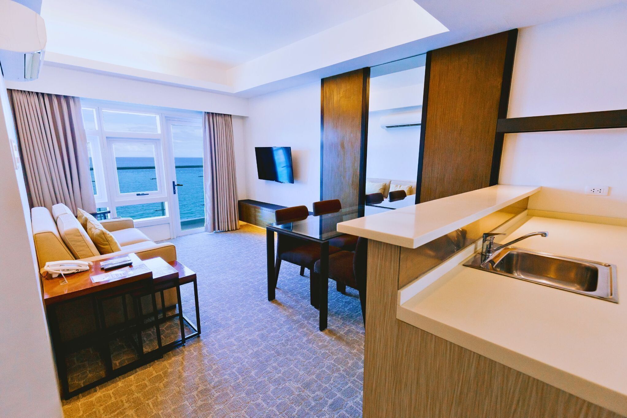 A hotel room with a living area, a dining table, a kitchen with a sink, and a TV mounted on the wall.