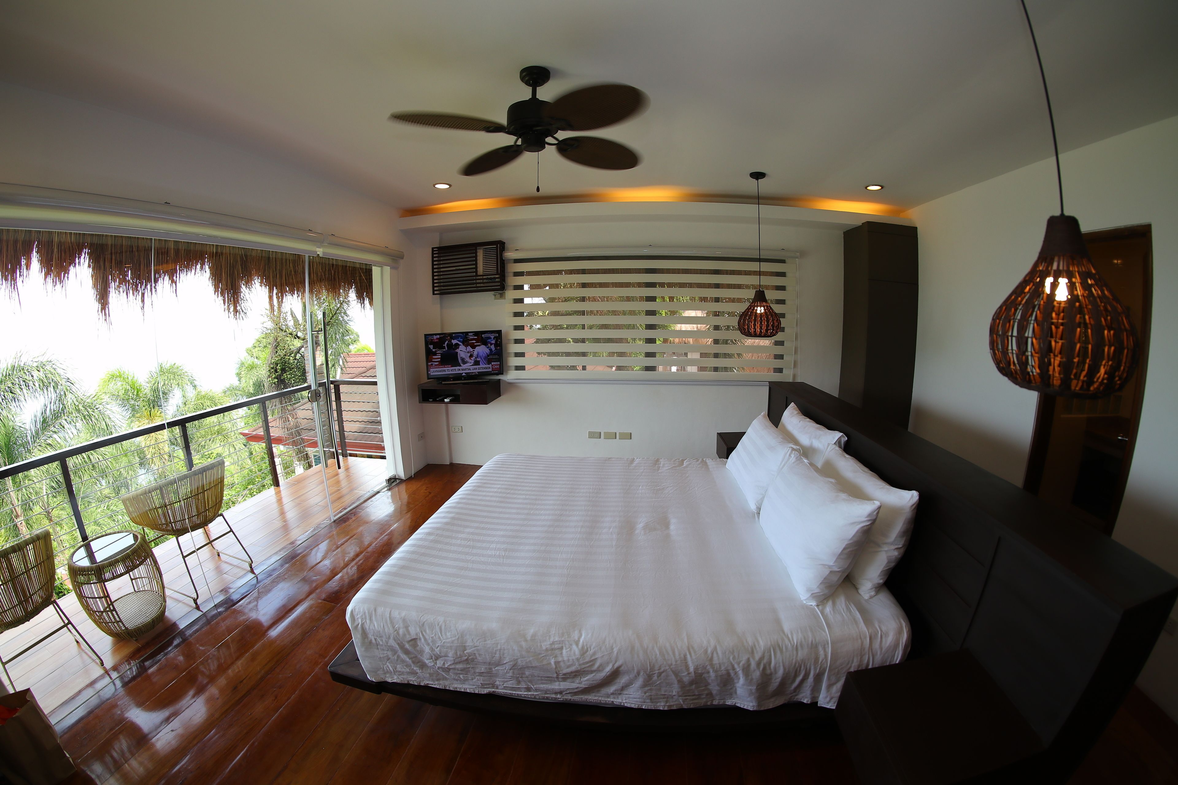 A bedroom with a large bed, a ceiling fan, a TV, and a balcony with chairs.