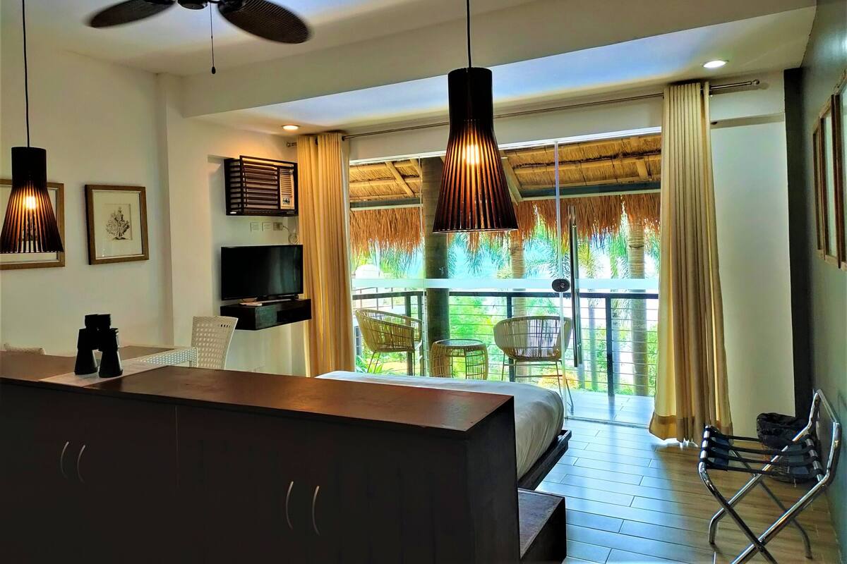A modern hotel room with a bed, a TV, a desk, and a balcony with a view of greenery.