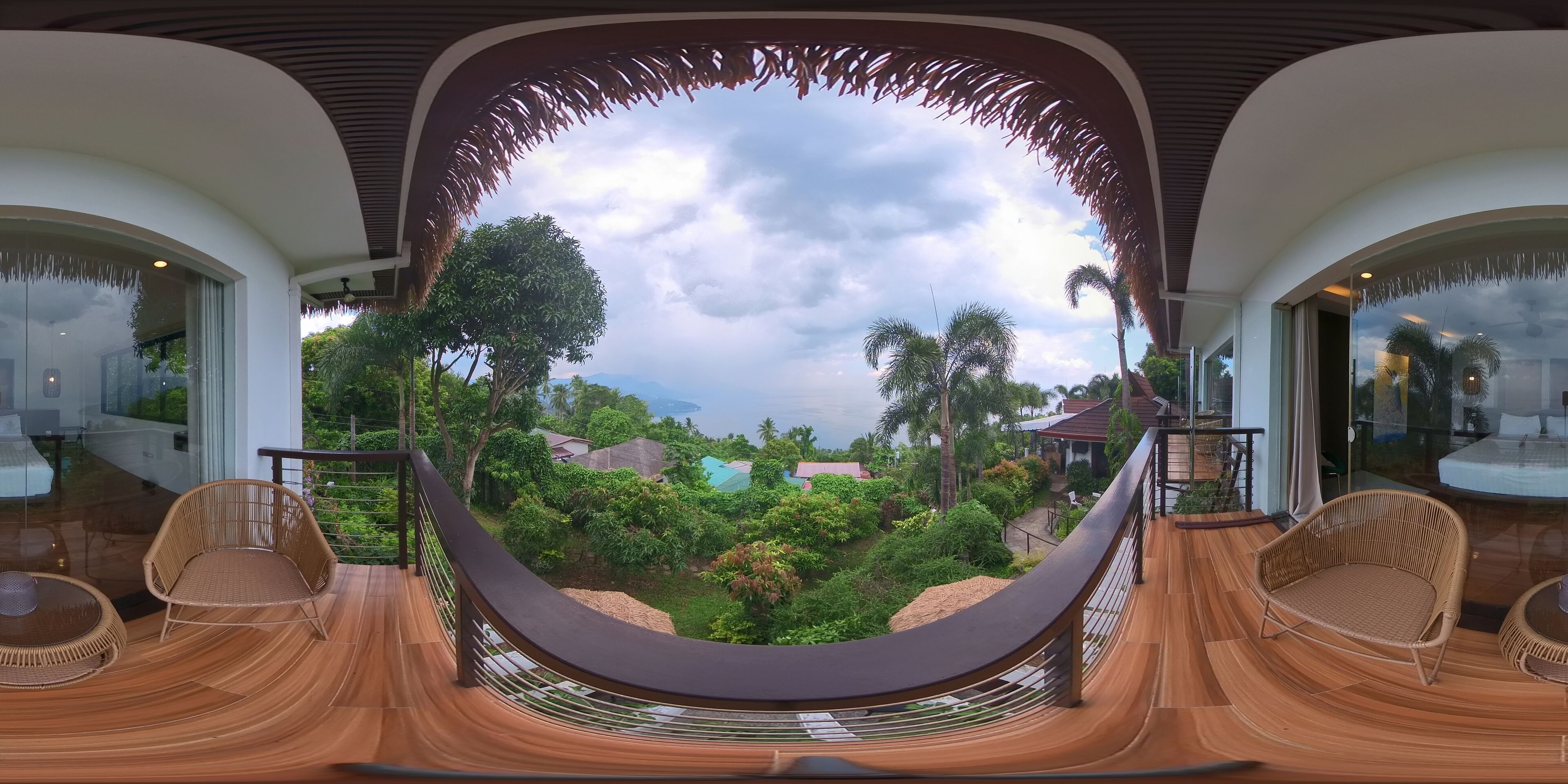A panoramic view from a balcony with wicker furniture and a glass railing, overlooking a lush garden and distant buildings.