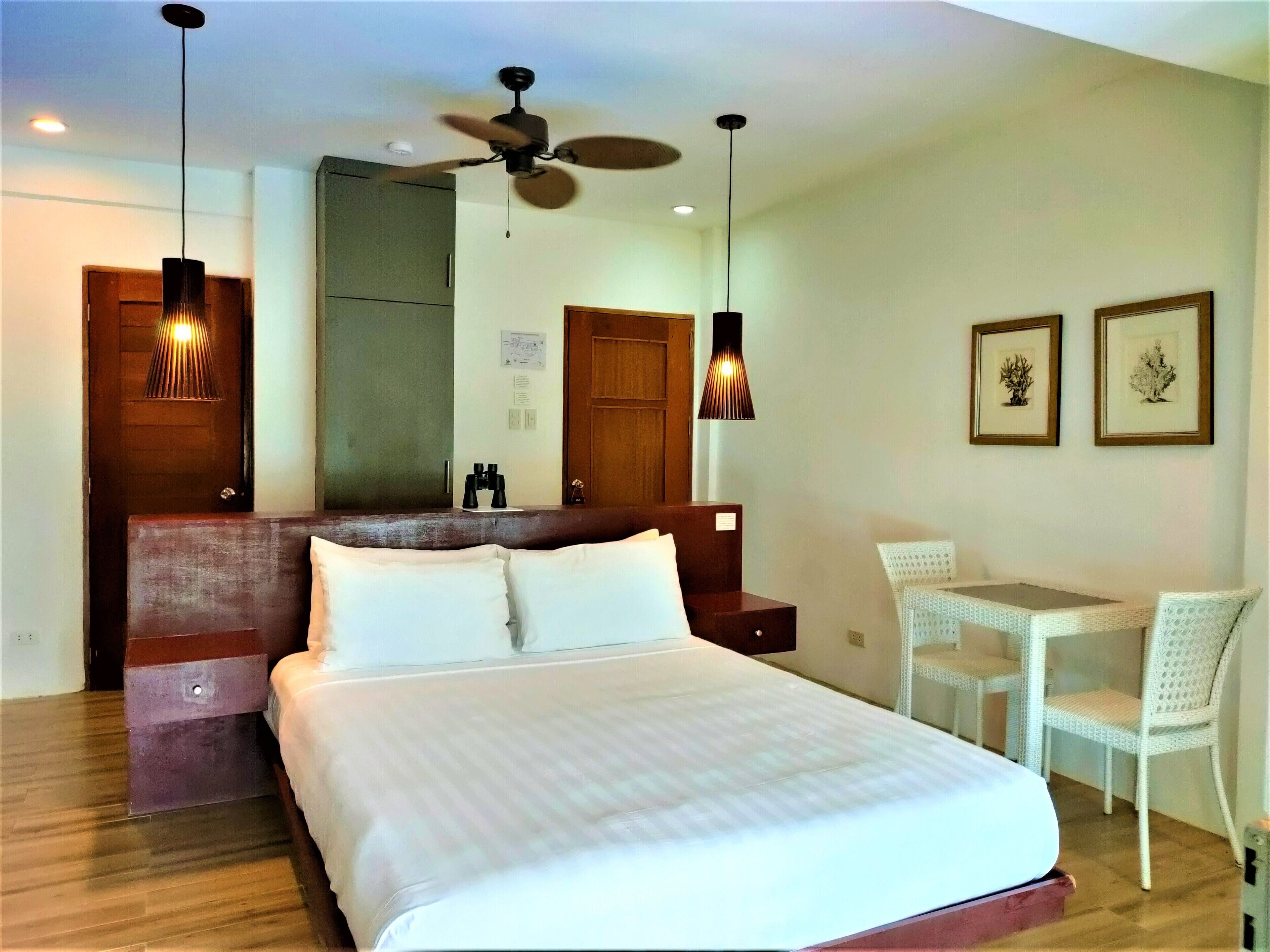 A hotel room with a large bed, a desk with a chair, a ceiling fan, and two framed pictures on the wall.