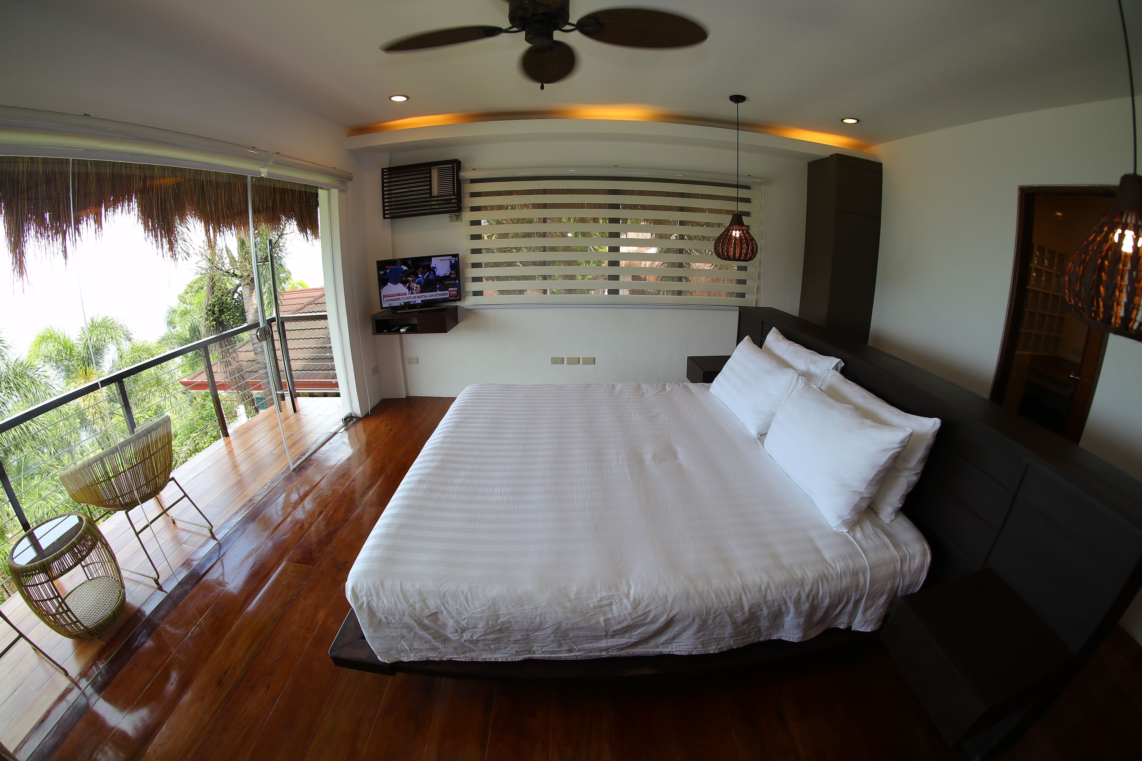 A bedroom with a large bed, a ceiling fan, a TV, and a balcony with chairs.