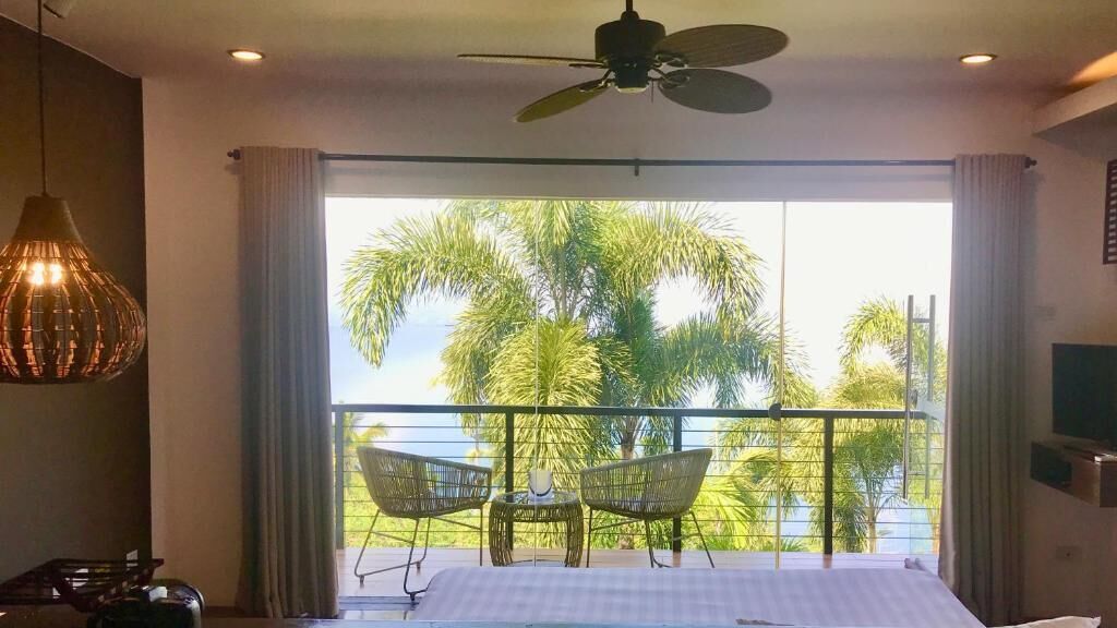 A bedroom with a bed, a ceiling fan, a table with chairs, and a balcony with a view of palm trees.