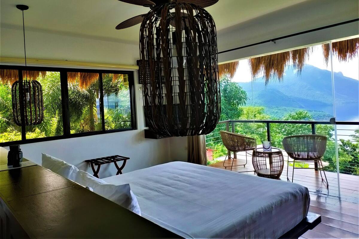 A bedroom with a large bed, a ceiling fan, and a view of lush greenery and mountains through large windows.