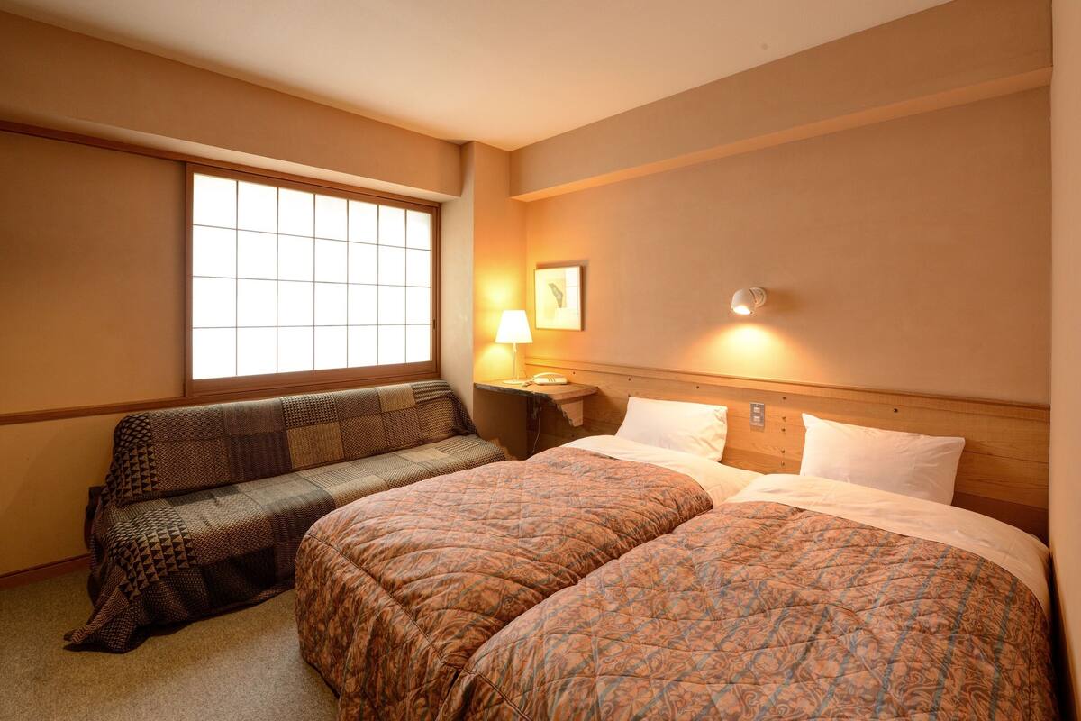 A hotel room with two beds, a window, a bedside table with a lamp, and a wall-mounted light.