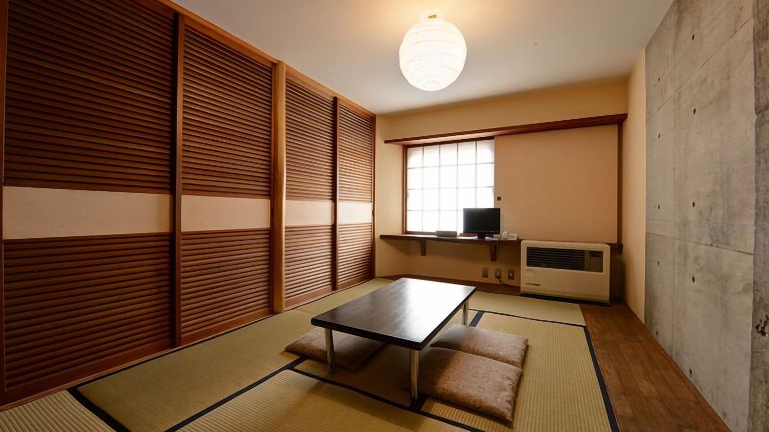 A traditional Japanese room with tatami flooring, a low table, cushions, sliding shoji doors, a television, and an air conditioning unit.