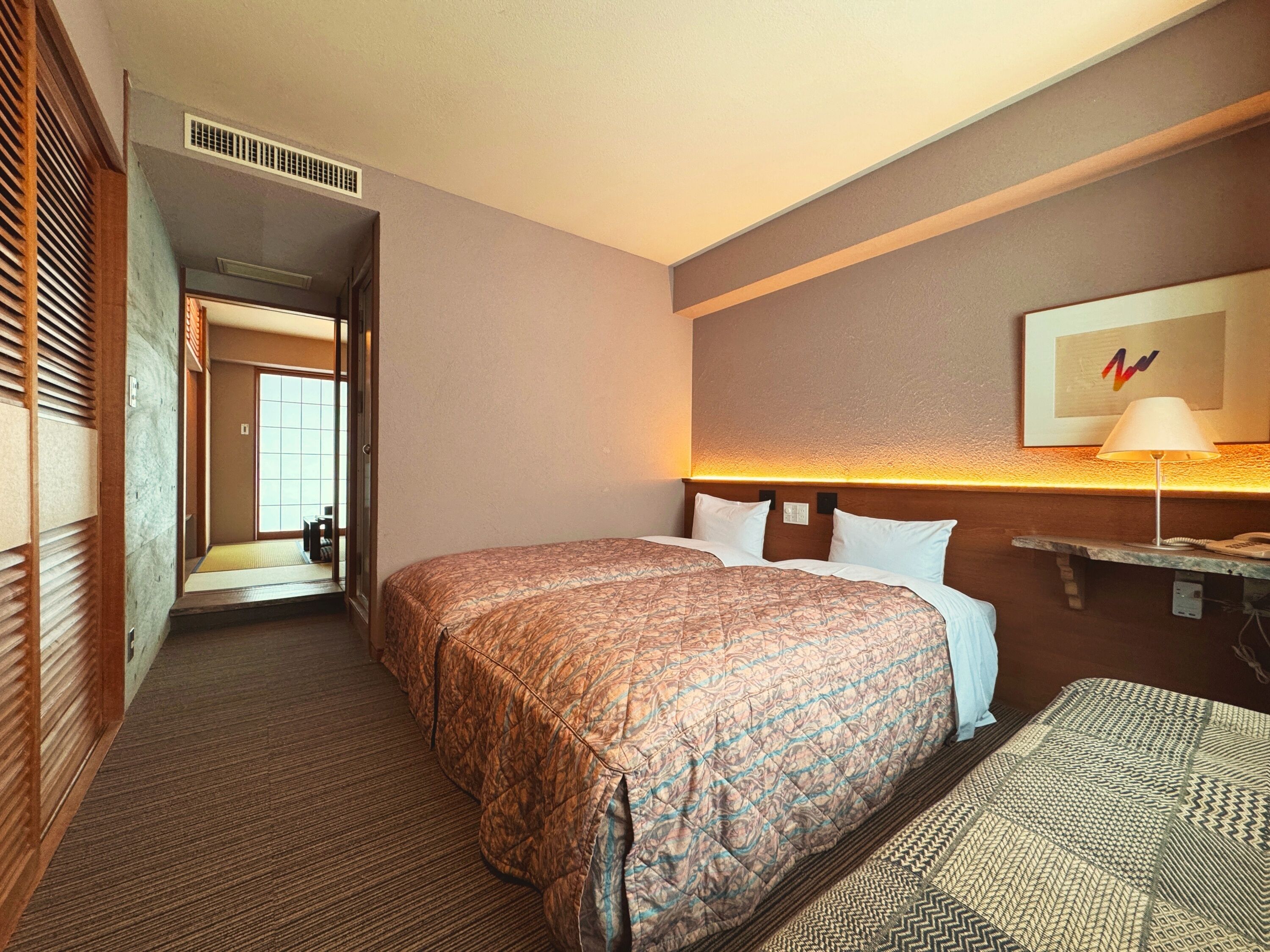 Una habitación de hotel con dos camas, una mesita de noche con lámpara, un televisor de pared y una puerta que da a otra habitación.