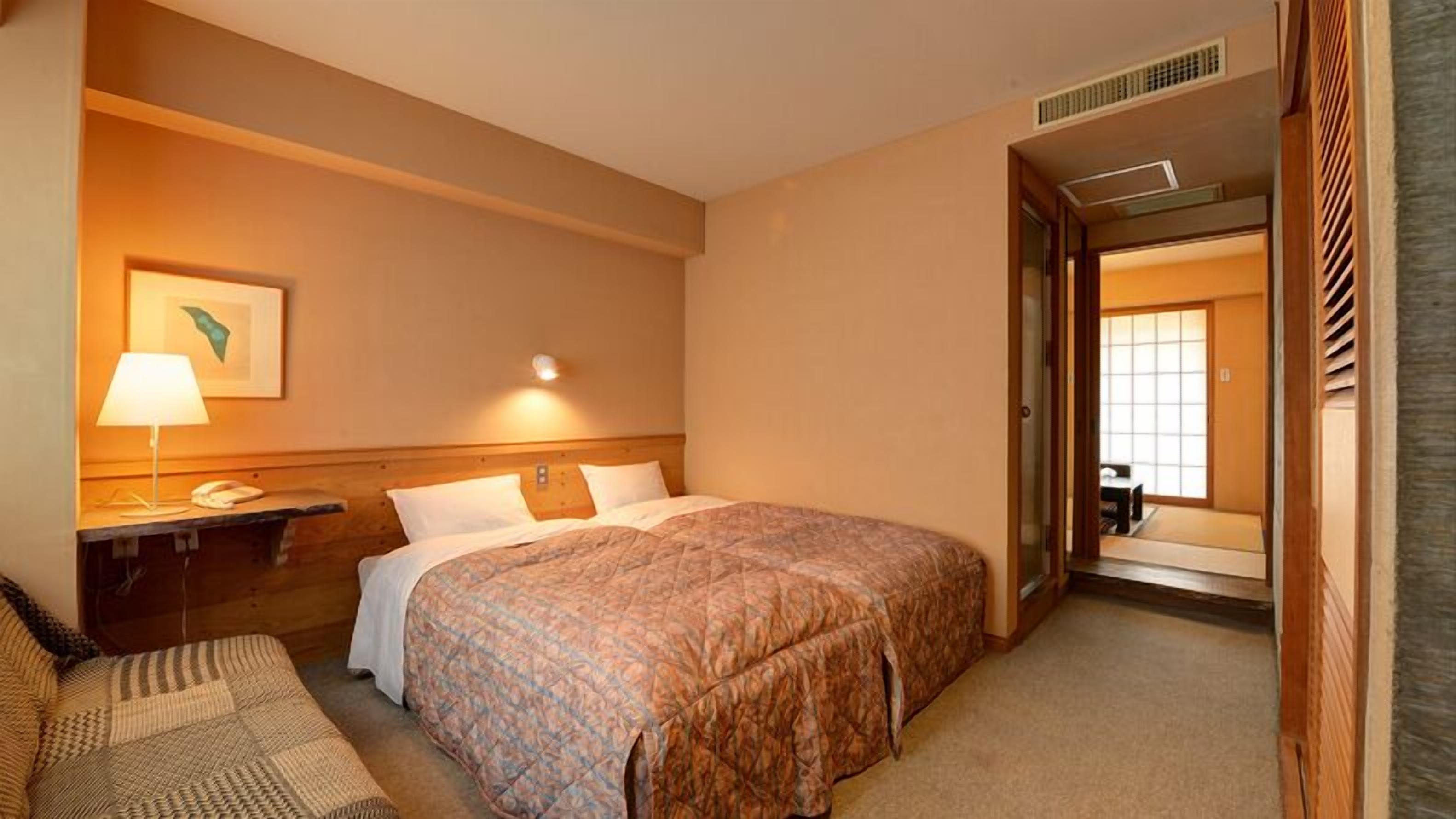 A hotel room with a bed, bedside table, lamp, and a view of another room through an open door.