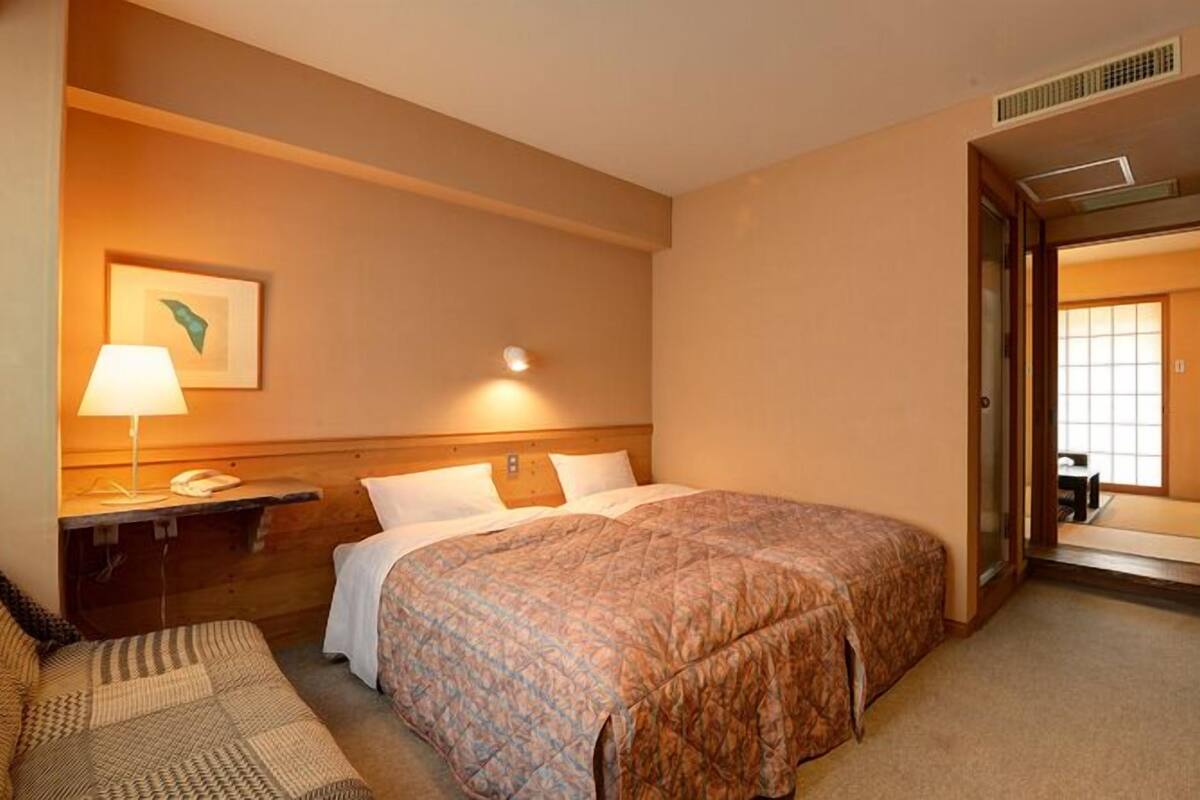 A hotel room with a bed, bedside table, lamp, and a view of another room through an open door.