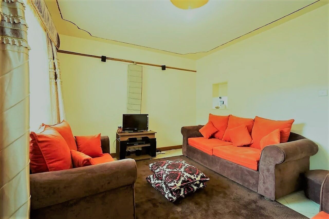 A living room with two sofas, one with orange cushions and another with brown cushions, a patterned ottoman, a television on a stand, and a window with curtains.