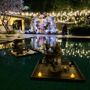 A nighttime outdoor setting with a reflective pool, decorative lights, and floral arrangements. - Estancia De Lorenzo Tarlac (Capas)