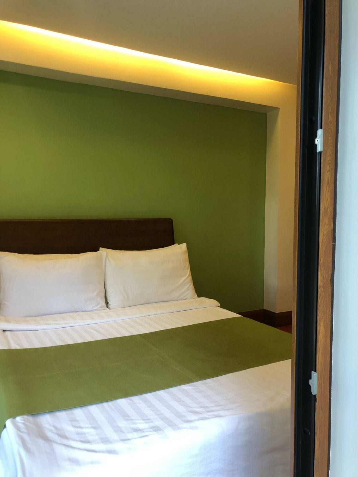 Ein ordentlich bezogenes Bett mit weißen Leinentüchern und einer grünen Tagesdecke, vor einer grünen Wand mit einem Holz-Kopfstück.