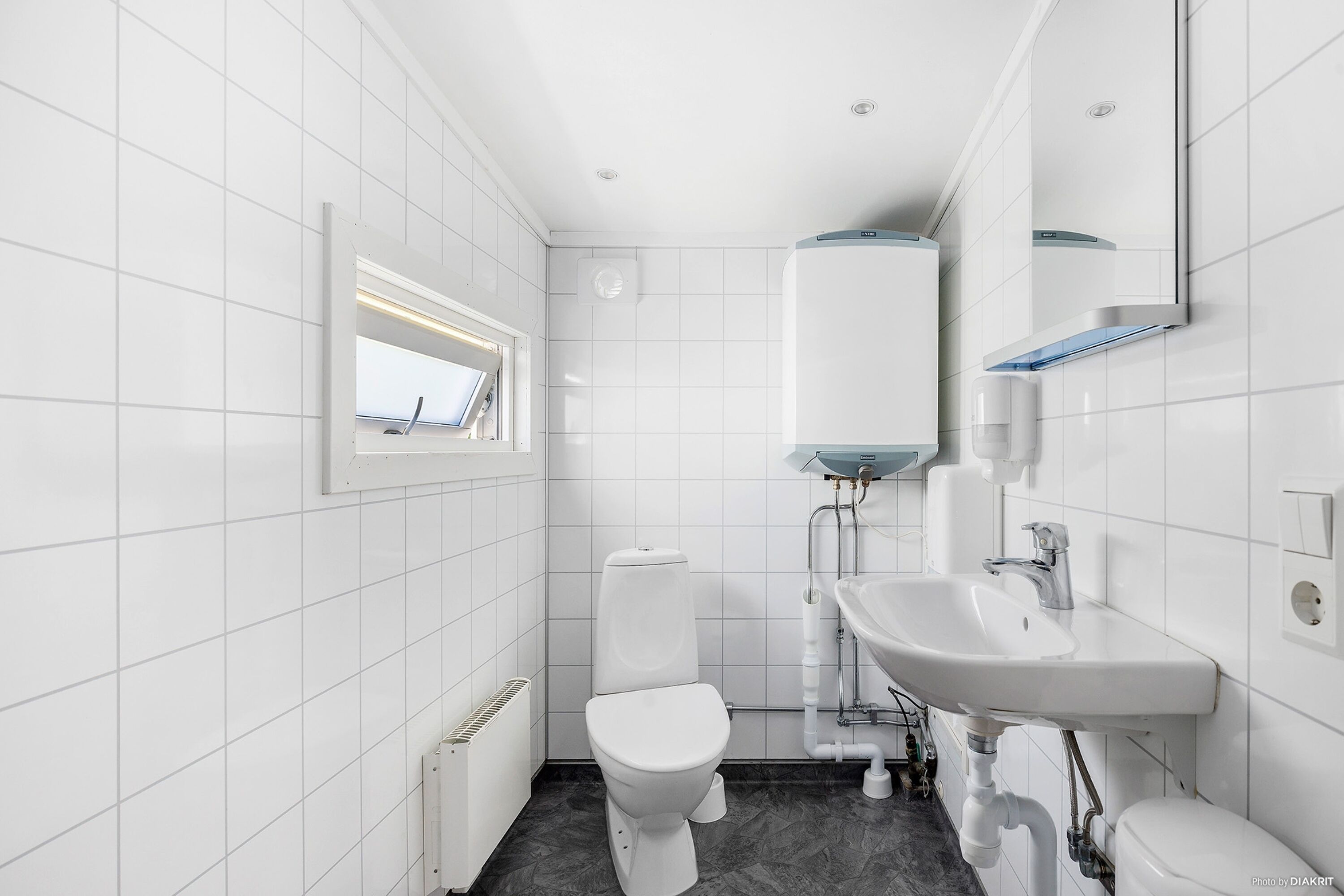 A modern bathroom with white tiles, a toilet, a sink, and a window.