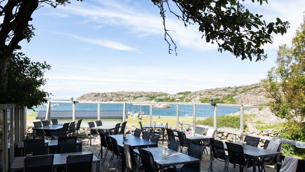 Un espace de restauration en plein air, avec des tables et des chaises, offrant une vue sur un paysage côtier composé de collines rocheuses et d’une étendue d’eau.