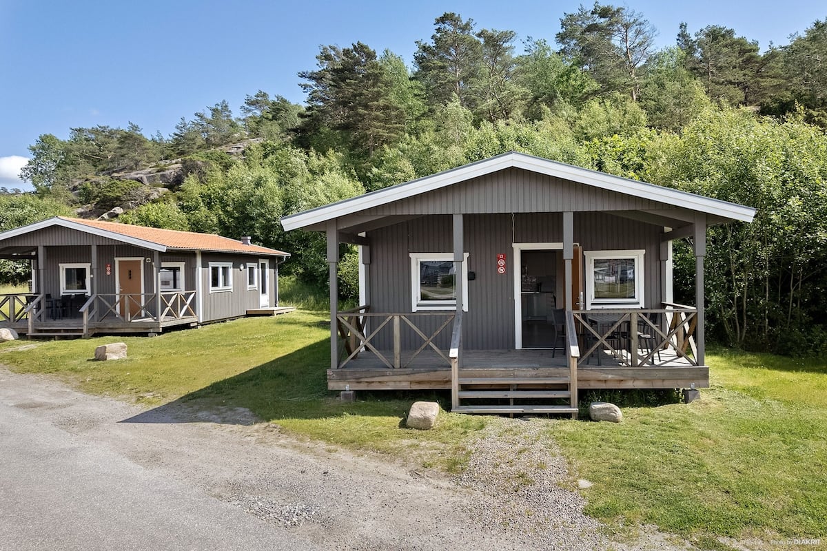 Two cabins with wooden decks and a porch, surrounded by greenery and a clear sky.