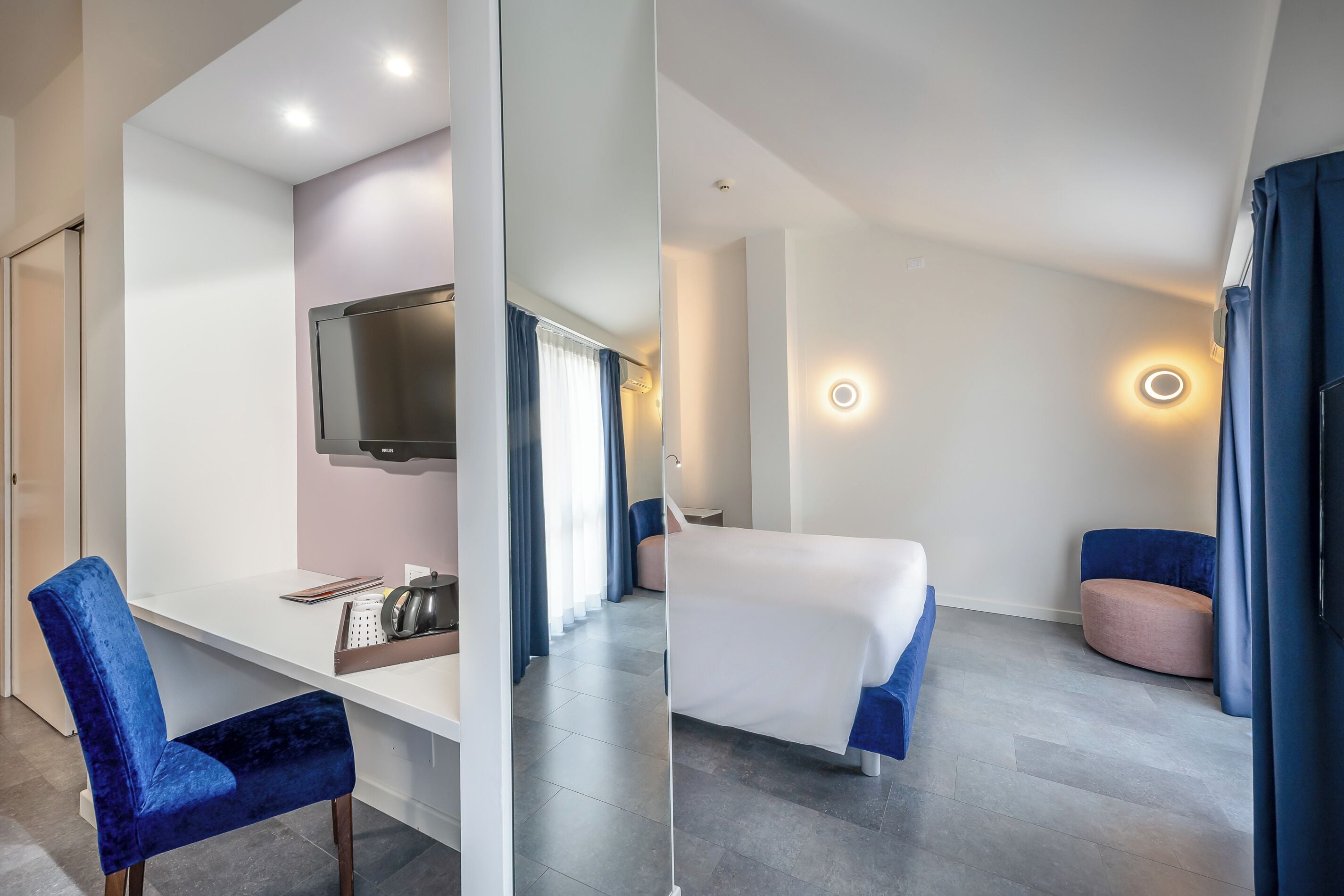 Moderni hotellihuone, jossa on taulutelevisio, työpöytä tuolilla, sänky ja oleskelualue, jossa on vaaleanpunainen tuoli.