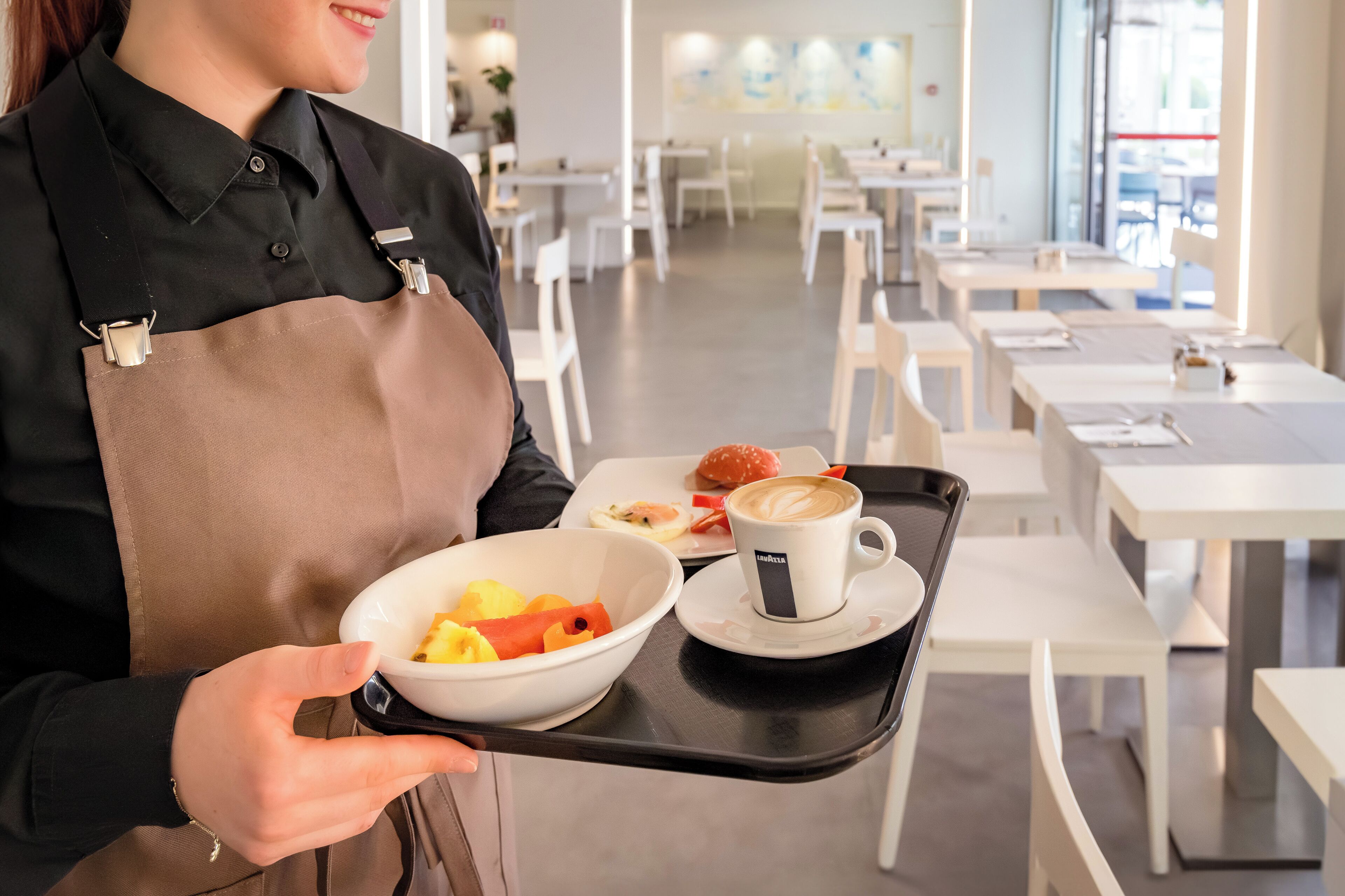 Eine Person hält ein Tablett mit einer Obstschale, einem Croissant auf einem Teller und einer Kaffeetasse in einem modernen Café.
