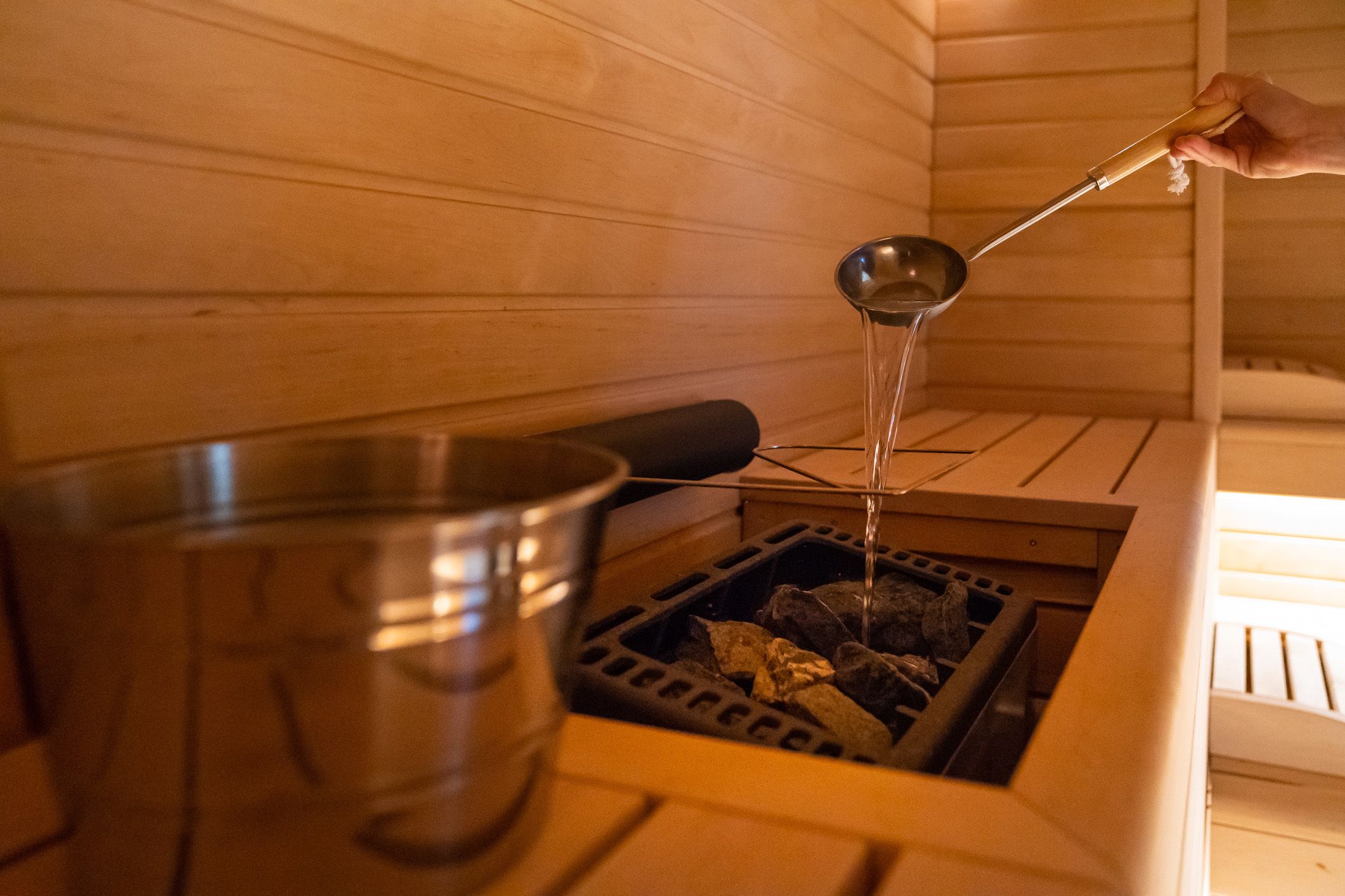 A sauna with a metal basin and a ladle pouring water into a stone-filled container.