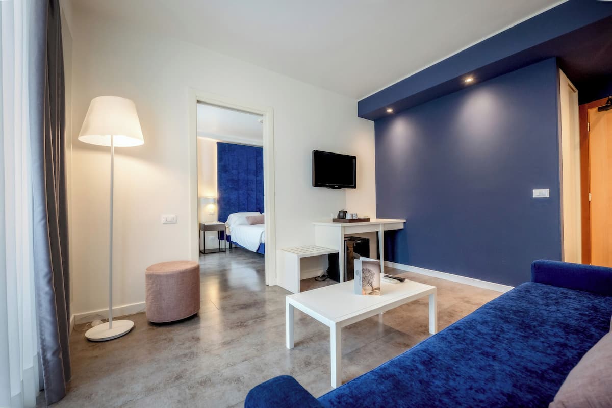 A modern hotel room with a blue accent wall, a flat-screen TV mounted above a desk, a white coffee table, and a blue sofa.