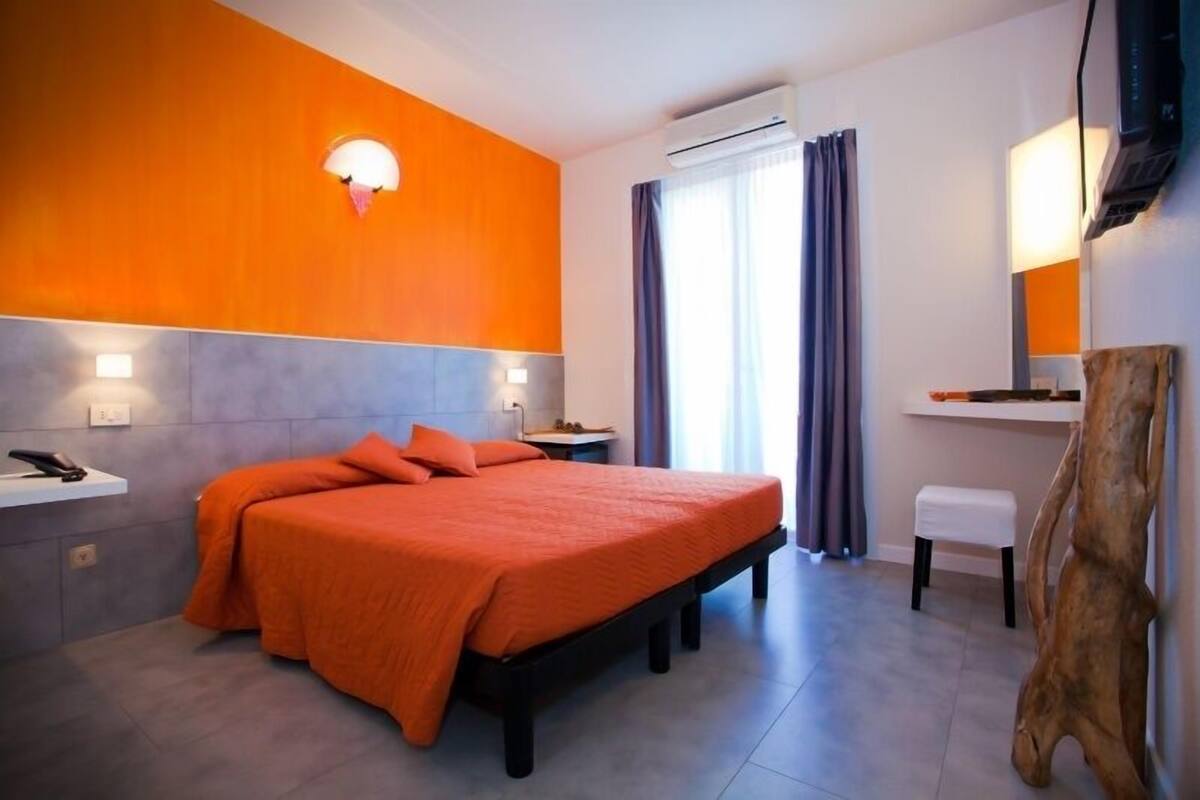 A modern hotel room with a large bed, a bedside table, a desk, and a chair.