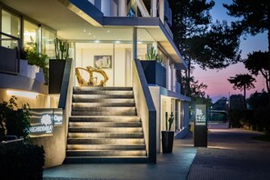 Un edificio moderno con una escalera que conduce a la entrada, rodeado de plantas en macetas y un cartel.