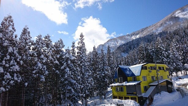 A yellow building with a blue roof surrounded by snow-covered trees and mountains in the background. - Allumeur (Minamiuonuma)