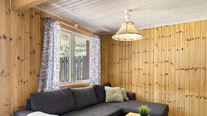 A cozy room with a wooden paneled wall, a grey sofa with cushions, a small wooden table with a plant, and a window with floral curtains. - First Camp Ekudden Mariestad (Mariestad)