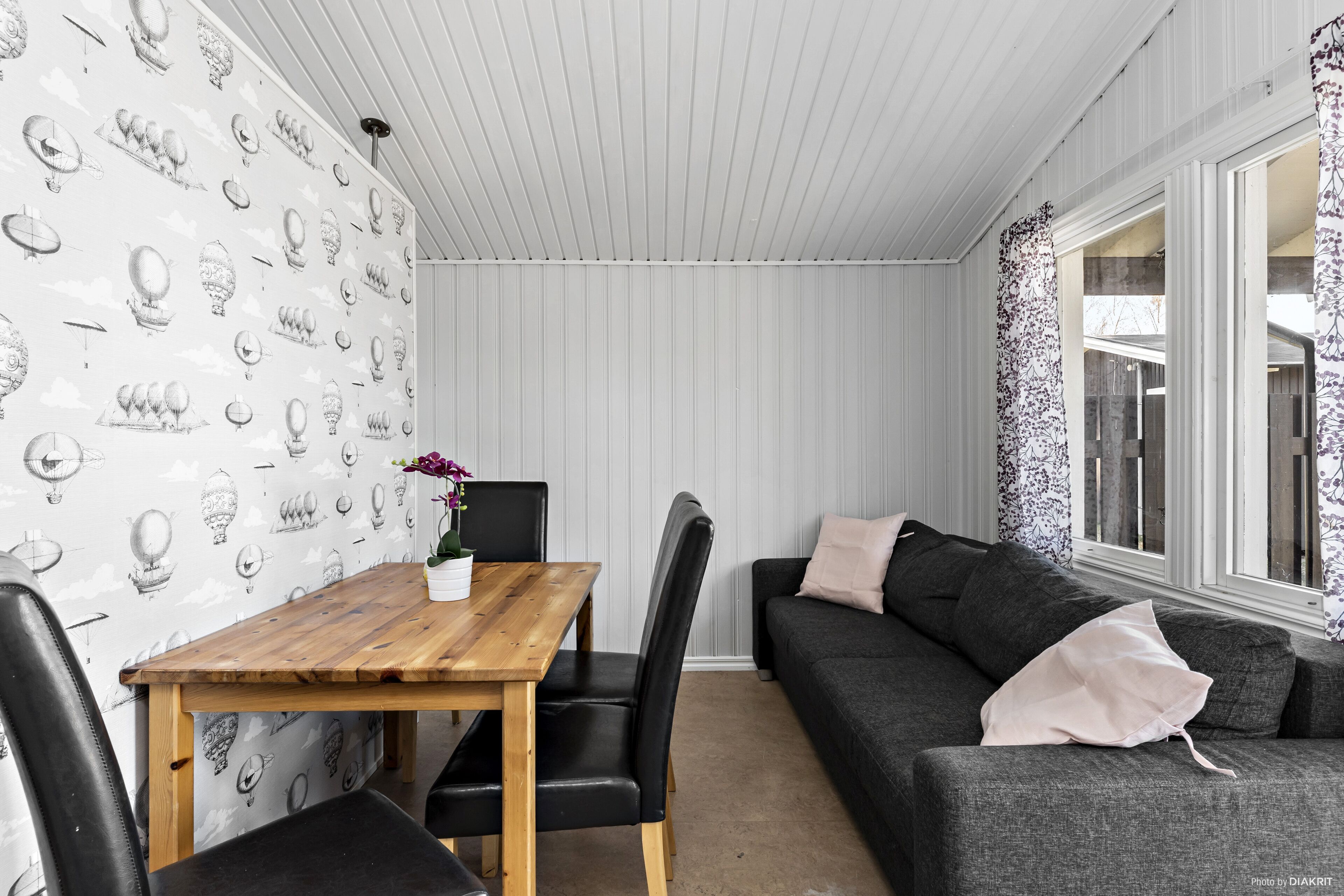 Quarto com mesa de jantar de madeira, cadeiras pretas, sofá cinza e janela com cortinas estampadas.