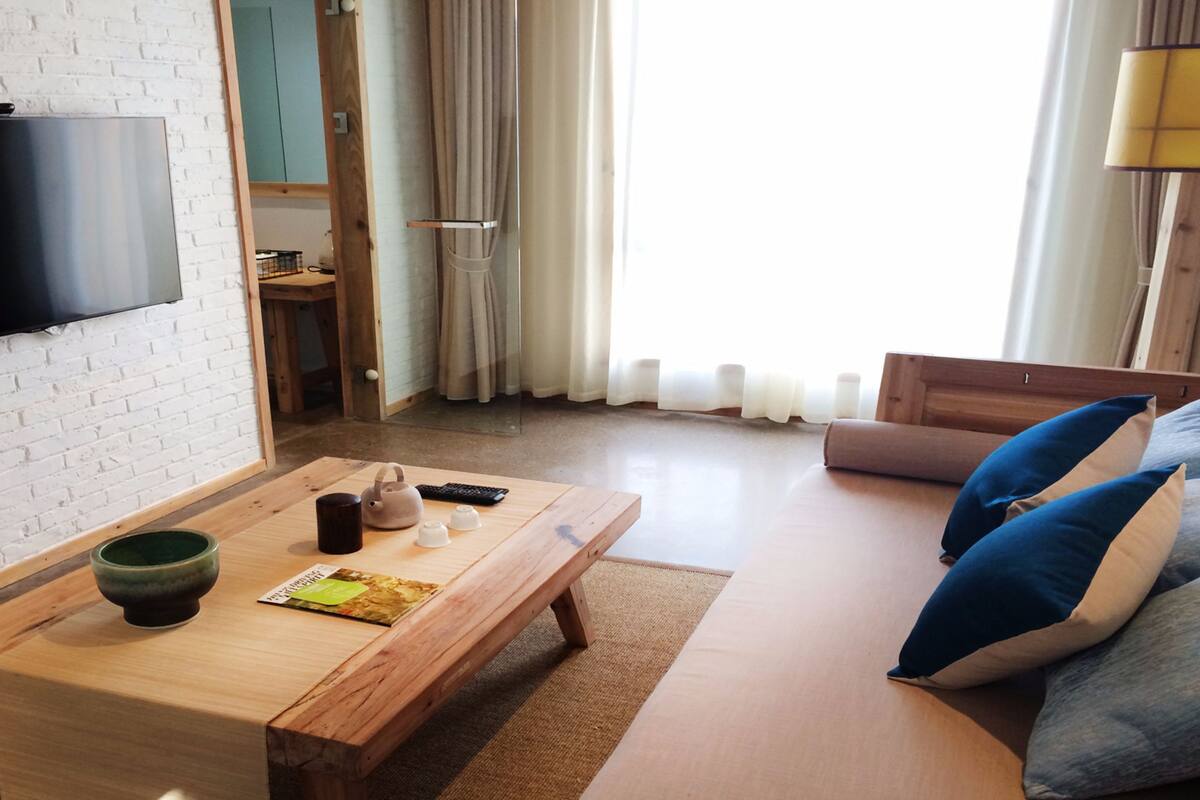 Una sala de estar moderna con una mesa de centro de madera, un sofá con cojines azules y beige, un televisor de pantalla plana montado en una pared de ladrillo blanco y vistas al exterior a través de grandes ventanales.