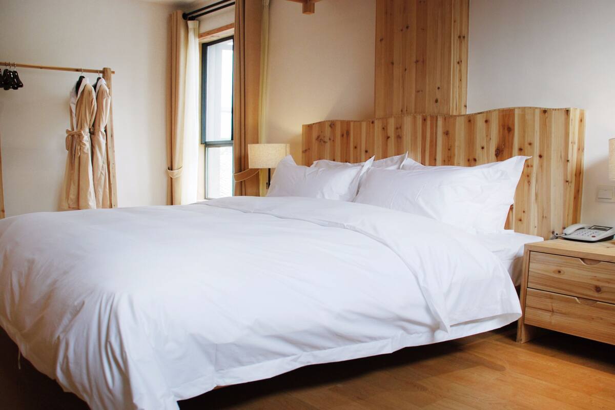 Una cama bien hecha en una habitación con paredes y suelo de madera. Hay una mesita de noche de madera con una lámpara y un teléfono encima.