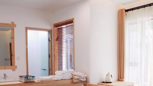 A modern hotel room with a sofa, a small wooden table, a mirror, a window with blinds, and a bathroom visible through an open door.