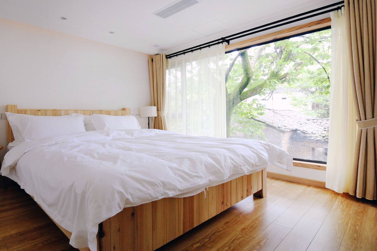 Un dormitorio con cama de madera, dos mesitas de noche, una ventana con cortinas y vistas al exterior con vegetación.