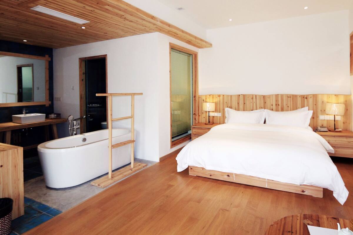 Un dormitorio moderno con suelo de madera, una cama grande, una mesita de noche con lámpara, una escalera de madera, un baño con bañera y un espejo.