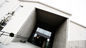 A modern building entrance with a concrete facade, large glass doors, and a small potted plant on the left. - Bryne Hotell (Time)