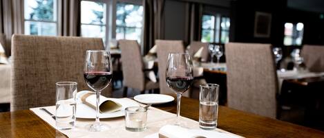A dining table set with wine glasses, water glasses, and plates in a restaurant with large windows and chairs.