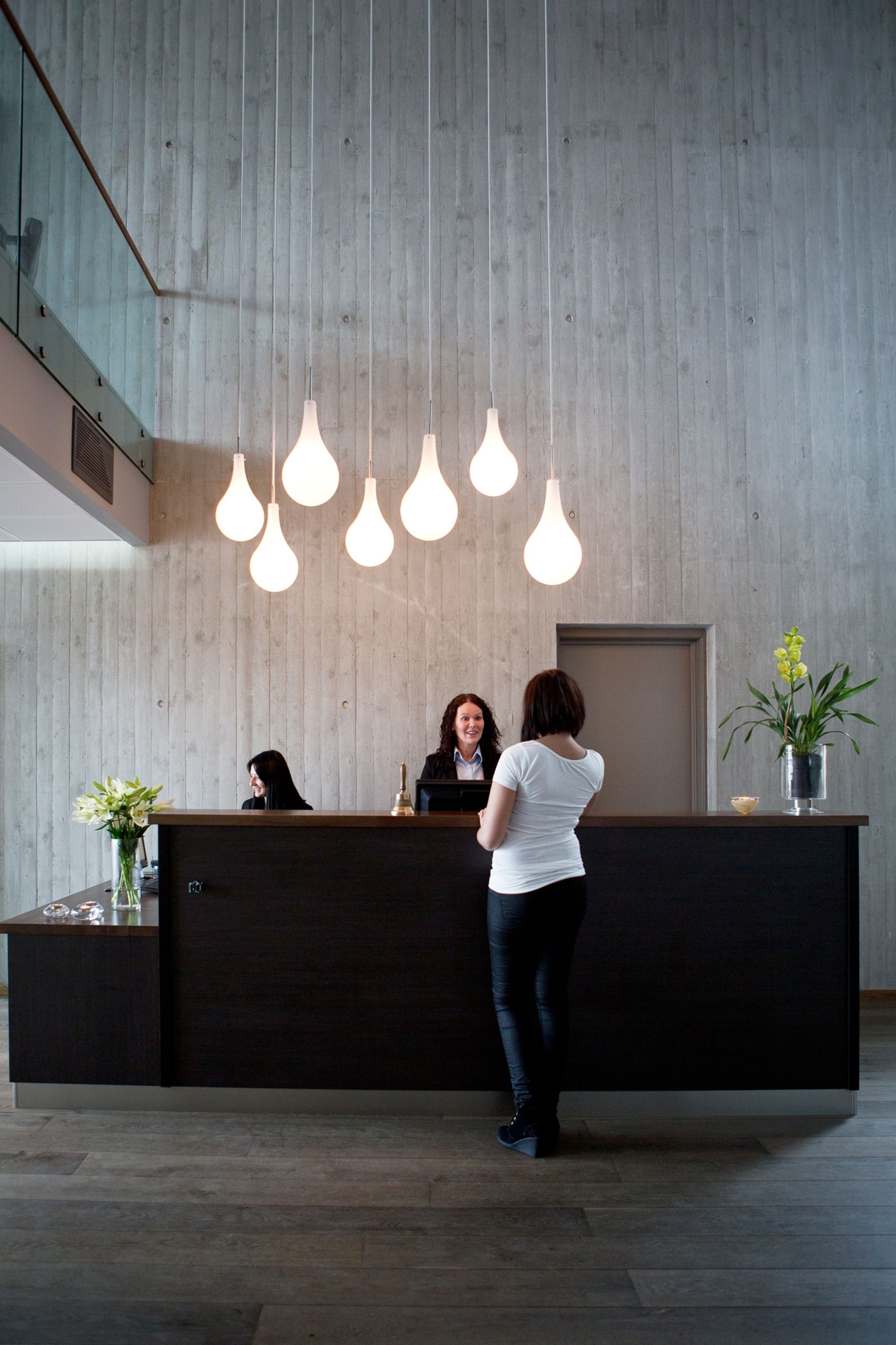 A reception desk with a modern design, featuring a dark wood finish and a glass partition. There are multiple light fixtures hanging from the ceiling, and a vase with flowers on the desk.