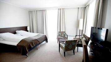 Habitación de hotel con dos camas, un escritorio con silla, televisor y ventanas amplias con cortinas.
