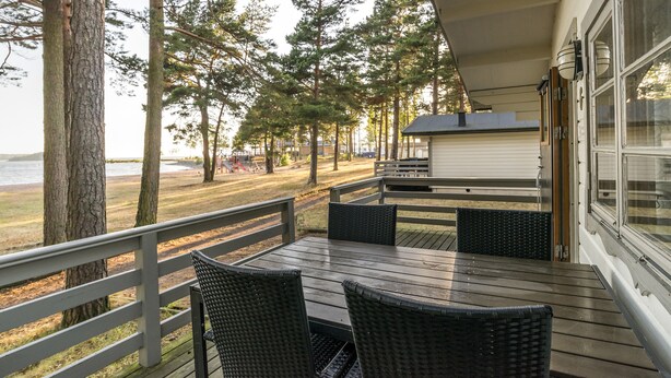 A covered outdoor deck with a table and chairs, overlooking a beach and forested area. - First Camp Mörudden Karlstad (Hammaro)