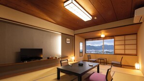 Una habitación japonesa tradicional con suelo de tatami, una mesa baja, sillas, un televisor y vistas a las montañas a través de amplias ventanas.