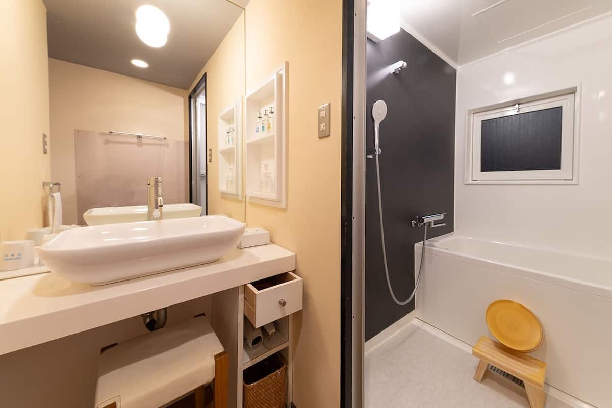 Un baño con lavamanos blanco, espejo, ducha y un pequeño taburete de madera.