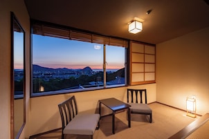 Habitación con un ventanal amplio que ofrece vistas a un paisaje urbano al atardecer. Hay dos sillas y una mesita.