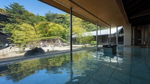 Una amplia piscina interior con paredes de vidrio transparente, techo de madera y vistas a un jardín ajardinado con árboles y rocas.