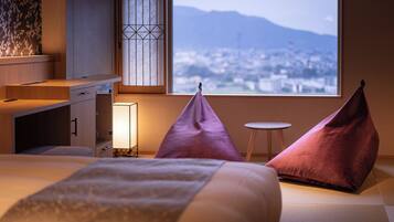 A hotel room with a bed, pillows, a bedside table, a lamp, and a view of a cityscape through a window.