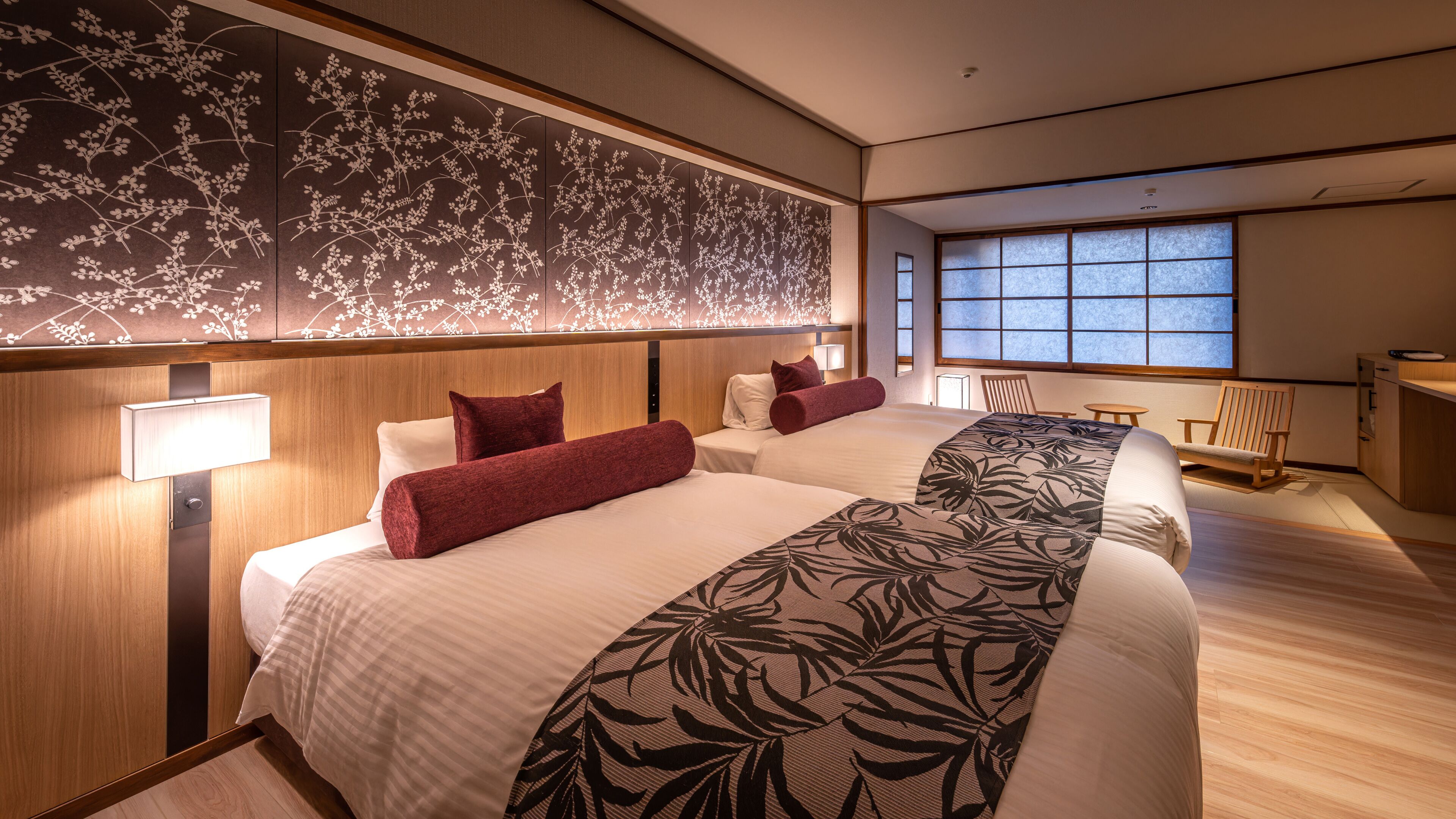 A hotel room with two beds, a large window, a wooden desk, and a chair.