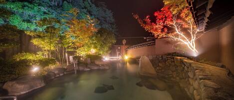 A serene nighttime setting with a stone-lined water feature, illuminated trees, and traditional architecture.