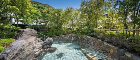 A hot spring surrounded by rocks and greenery, with a stone wall and a metal railing.