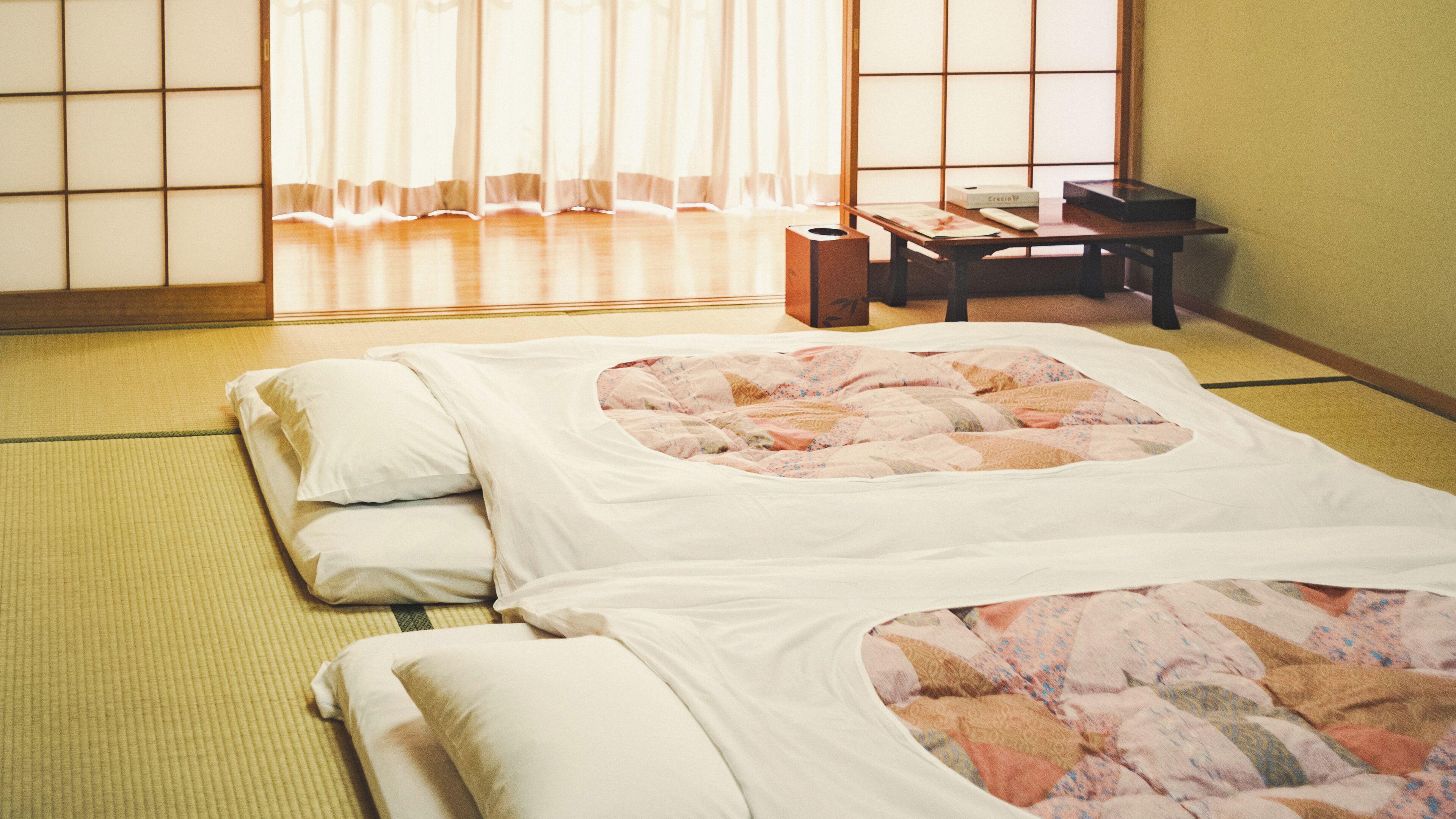 A traditional Japanese tatami mat room with a bed, pillows, a desk, and a window with shoji screens.