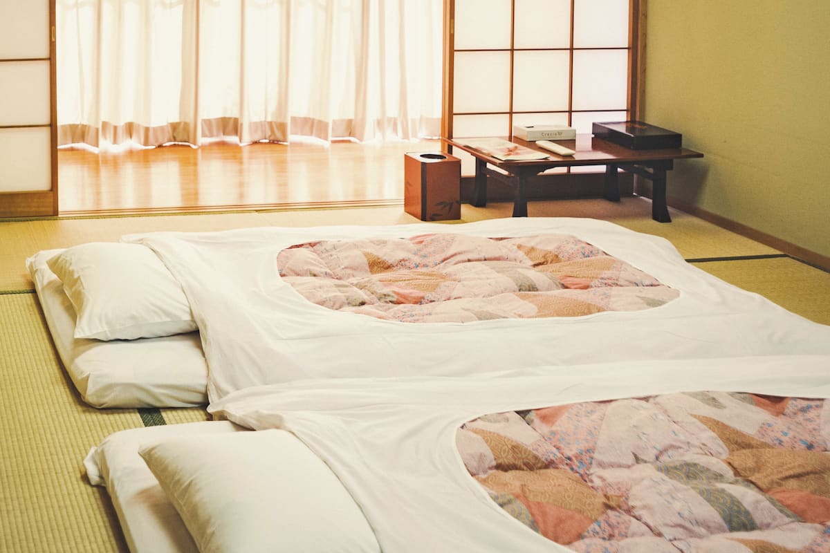 Una habitación tradicional japonesa con esteras de tatami, una cama, almohadas, un escritorio y una ventana con paneles shoji.