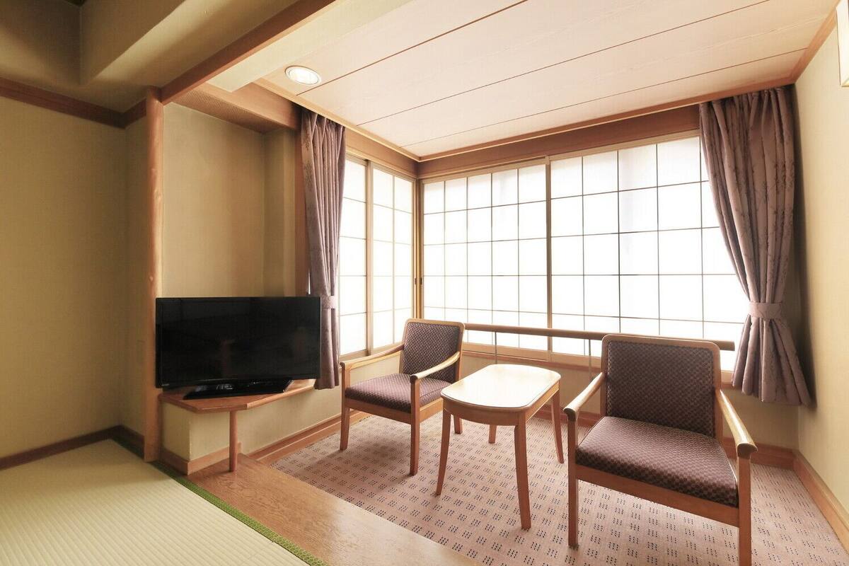 Una habitación con suelo de tatami, un televisor sobre un mueble bajo, dos sillones cómodos, una mesita y ventanas grandes con cortinas.