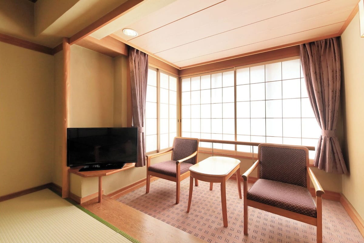 Habitación de hotel con suelo de tatami, televisión, sofá, mesa y ventanas amplias con cortinas.